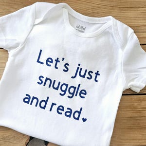 May include: A white short-sleeved baby bodysuit featuring the dark blue printed text "Let's just snuggle and read." with a small heart icon at the end of the sentence. The garment has a round neckline and a "child of mine" label printed inside.