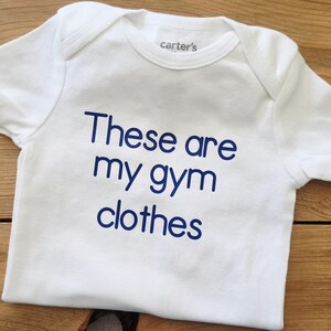May include: A white short-sleeved bodysuit with a crew neckline and the text "These are my gym clothes" printed in dark blue lettering on the front. The garment features a Carter's brand label on the inside of the neck area and short sleeves.