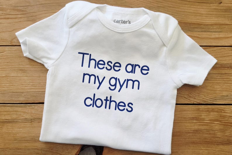 May include: A white short-sleeved bodysuit with a crew neckline and the text "These are my gym clothes" printed in dark blue lettering on the front. The garment features a Carter's brand label on the inside of the neck area and short sleeves.