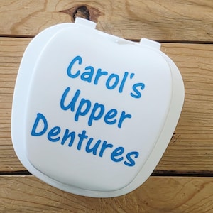 May include: A white denture case with the words "Carol's Upper Dentures" in blue script. The case is rectangular with rounded edges and a hinged lid. The case is sitting on a wooden surface.
