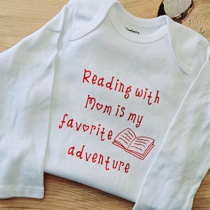 May include: White long-sleeved baby onesie with red text that reads "Reading with Mom is my favourite adventure" and a red illustration of an open book.