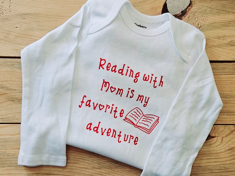 May include: White long-sleeved baby onesie with red text that reads "Reading with Mom is my favourite adventure" and a red illustration of an open book.