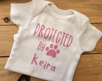 May include: White baby onesie with pink text that reads "PROTECTED BY Keira" and a pink paw print.