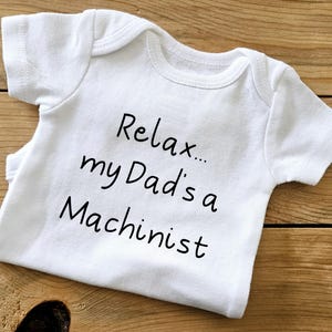 May include: White baby onesie with black text that reads "Relax... my Dad's a Machinist".