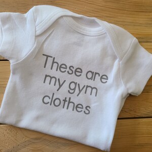 May include: A white short-sleeved baby bodysuit featuring the grey printed text "These are my gym clothes" centered on the front. The garment has a round neckline and short sleeves, constructed with a standard envelope-style shoulder design.