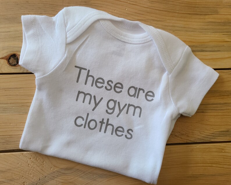 May include: A white short-sleeved baby bodysuit featuring the grey printed text "These are my gym clothes" centered on the front. The garment has a round neckline and short sleeves, constructed with a standard envelope-style shoulder design.