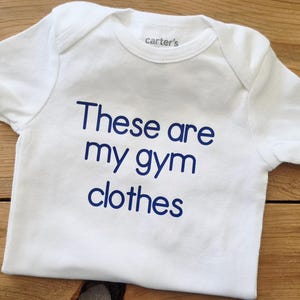 May include: A white short-sleeved bodysuit with a crew neckline and the text "These are my gym clothes" printed in dark blue lettering on the front. The garment features a Carter's brand label on the inside of the neck area and short sleeves.