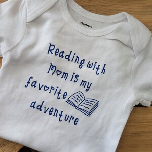 May include: White baby onesie with blue text that reads "Reading with Mom is my favourite adventure" and a blue illustration of an open book.