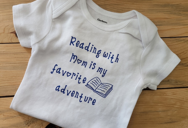 May include: White baby onesie with blue text that reads "Reading with Mom is my favourite adventure" and a blue illustration of an open book.