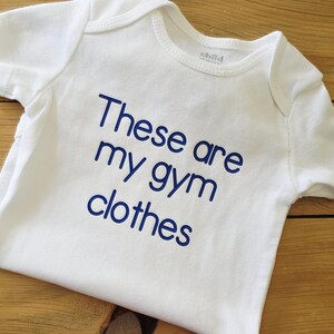 May include: A white short-sleeved baby bodysuit featuring the dark blue text "These are my gym clothes" printed on the front. The garment has a round neckline and short sleeves, with a simple design focused on the bold, sans-serif lettering.