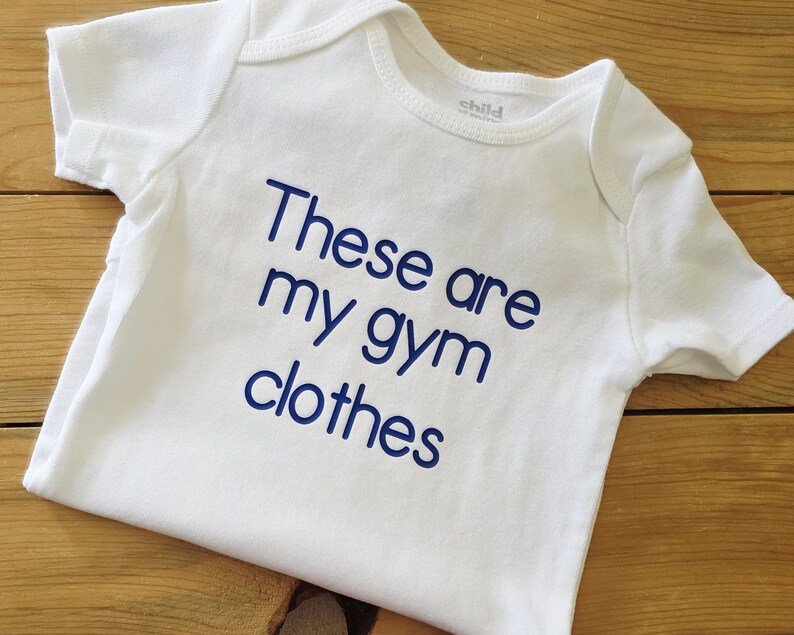 May include: A white short-sleeved baby bodysuit featuring the dark blue text "These are my gym clothes" printed on the front. The garment has a round neckline and short sleeves, with a simple design focused on the bold, sans-serif lettering.