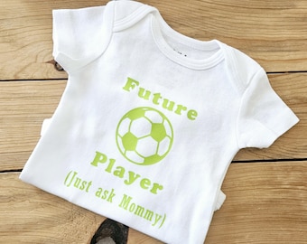 May include: White baby onesie with green text that reads "Future Player (Just ask Mommy)" and a green soccer ball graphic.
