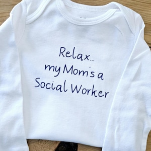 May include: White baby onesie with long sleeves. The text "Relax... my Mom's a Social Worker" is written in blue script across the front. The onesie is laying on a wooden surface.