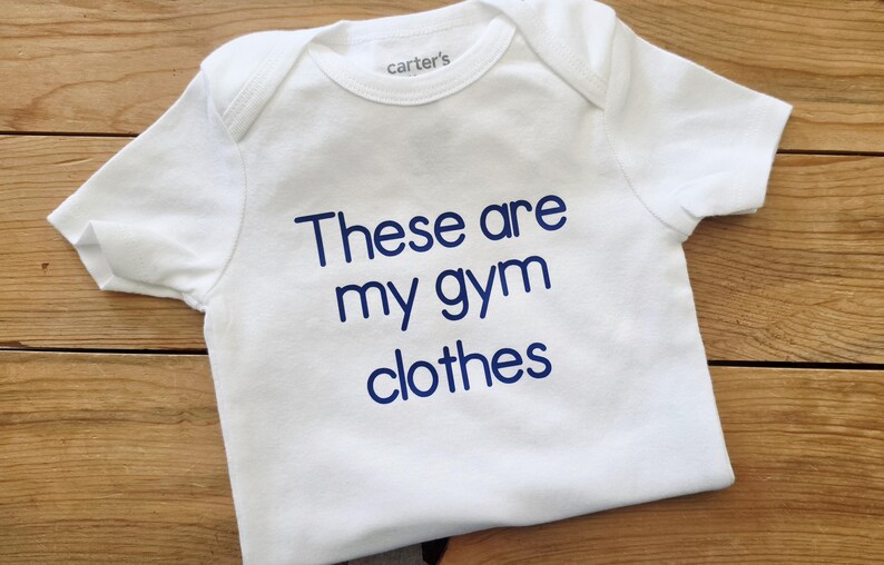 May include: A white short-sleeved bodysuit with a crew neckline and the text "These are my gym clothes" printed in blue on the front. The garment features a "carter's" brand label on the inside of the collar and has a simple, clean design.