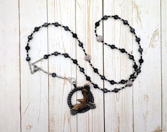 May include: A necklace with black and grey speckled beads on a black cord. The pendant is a circular design with a bronze-coloured eagle in flight. The necklace has silver-coloured metal accents.