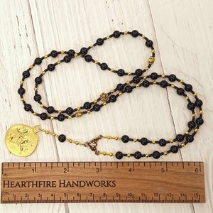 May include: A black and gold beaded necklace with a gold pendant featuring a figure of a person on horseback. The necklace is 15 inches long and is shown on a wooden surface with a ruler.