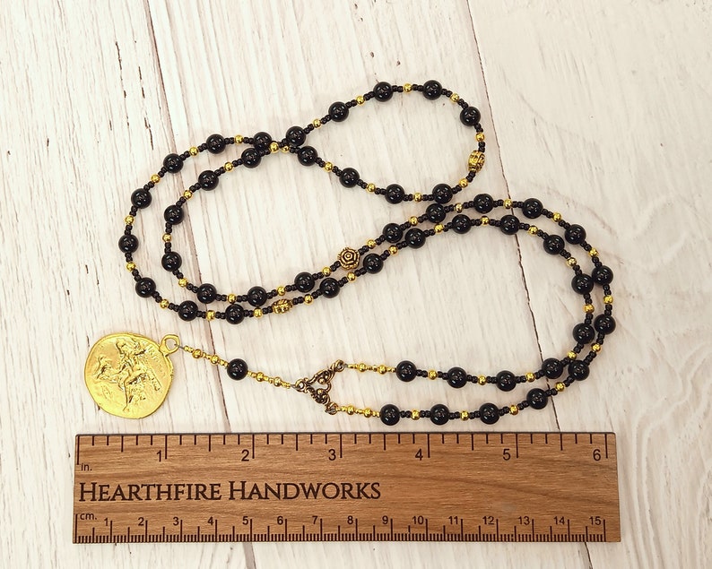 May include: A black and gold beaded necklace with a gold pendant featuring a figure of a person on horseback. The necklace is 15 inches long and is shown on a wooden surface with a ruler.