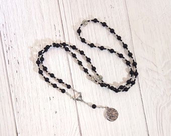 May include: A black beaded rosary necklace with silver-toned detailing. The necklace has round black beads and decorative silver-toned connectors. A silver-toned pendant with a detailed design hangs at the bottom.