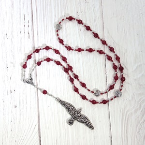 Puede incluir: Un rosario de cuentas rojas con detalles plateados y un colgante en forma de pájaro. Las cuentas son de color rojo intenso y el rosario se exhibe sobre una superficie de madera blanca.