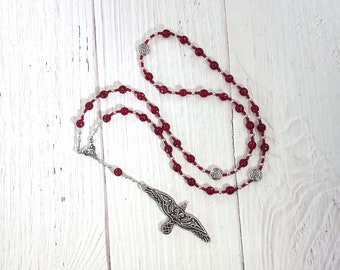 May include: A red beaded rosary with silver-toned accents and a detailed pendant in the shape of a bird. The beads are a deep red color, and the rosary is displayed on a white wooden surface.