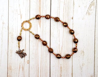 May include: A beaded necklace featuring faceted metallic bronze beads interspersed with smaller gold-toned seed beads. The necklace includes a circular twisted metal clasp and a bronze-toned charm shaped like a stylized animal head with pointed ears.