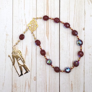 Op de afbeelding: Een rozenkrans met facetgeslepen, dieprode kralen en goudkleurige accenten. De kralen zijn geregen aan een rood koord. Een goudkleurige hanger van een figuur met een staf hangt aan de rozenkrans. De achtergrond is een lichte houtnerf.