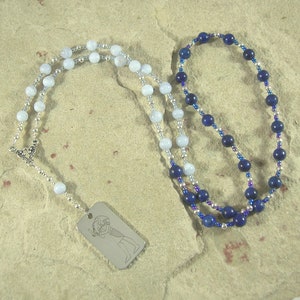 May include: Two necklaces with silver chain and beads. One necklace has light blue beads and a silver tag with an engraved image. The other necklace has dark blue beads with silver accents.