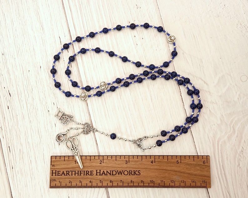 Puede incluir: Un collar de cuentas azules y plateadas con un dije plateado de un par de tijeras y una carretilla. El collar mide 38 cm de largo.