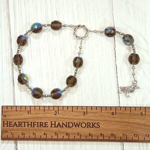 May include: A brown crystal beaded mala with a silver crow charm. The mala is approximately 4 inches long and is laid out on a wooden ruler with the text "HEARTHFIRE HANDWORKS" printed on it.