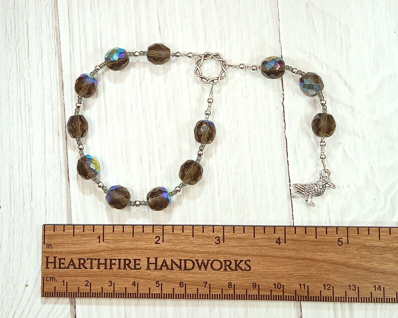 May include: A brown crystal beaded mala with a silver crow charm. The mala is approximately 4 inches long and is laid out on a wooden ruler with the text "HEARTHFIRE HANDWORKS" printed on it.