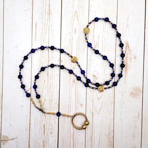 May include: A rosary necklace featuring dark blue and black beads with gold-colored accents and decorative medallions. The necklace is laid out on a light-colored wooden surface.