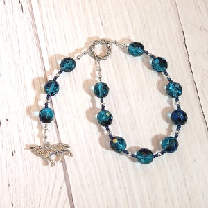 Puede incluir: Un collar de cuentas de cristal azul turquesa con un dije de lobo plateado. El collar está enhebrado en una cadena plateada.