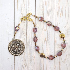 May include: A handcrafted rosary featuring faceted purple beads, gold-colored accents, and a Celtic knot pendant. The rosary has a decorative gold-colored top and is displayed on a light wood surface.
