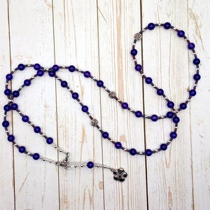 May include: A rosary with deep purple beads and silver-toned accents. The beads are connected by a silver chain, with decorative silver rose and flower charms. The rosary is displayed on a light wood-grain surface.