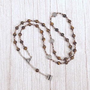May include: A rosary necklace featuring tiger eye beads and silver-toned metal accents. The beads are a mix of brown and gold, strung together with smaller brown beads. The necklace includes decorative Celtic knot charms and a cross pendant.