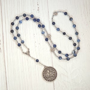 May include: A silver necklace with a large silver pendant featuring a circular design with runes. The necklace is made with blue and silver beads.