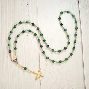 May include: A green and gold beaded necklace with a gold arrow charm. The necklace is made of small, round beads and is strung on a gold chain.