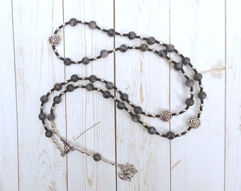 May include: A black and silver beaded rosary with a Celtic knot design. The rosary is made with dark grey beads and silver accents.