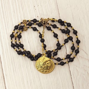 May include: A black onyx beaded necklace with a gold pendant featuring a profile of a person. The necklace has a gold clasp and is layered with two strands of beads.
