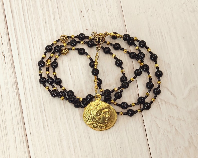 May include: A black onyx beaded necklace with a gold pendant featuring a profile of a person. The necklace has a gold clasp and is layered with two strands of beads.