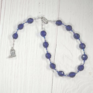 Może przedstawiać: Naszyjnik z ciemnoniebieskich koralików kryształowych z srebrnym zawieszką w kształcie drzwi. Naszyjnik wykonany jest z fasetowanych koralików i srebrnego łańcuszka.