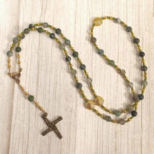 May include: A rosary with olive green and gray beads, connected by gold-colored chain. The rosary features a decorative cross pendant and gold-colored Celtic knot accents. The beads are approximately 0.25 inches in diameter.
