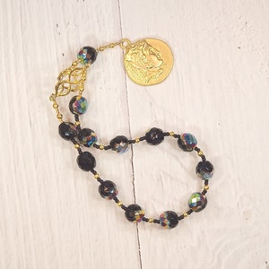 Pode incluir: Um bracelete de contas pretas com um fecho dourado que apresenta o perfil de uma pessoa. As contas s&atilde;o facetadas e t&ecirc;m um brilho iridescente arco-&iacute;ris.