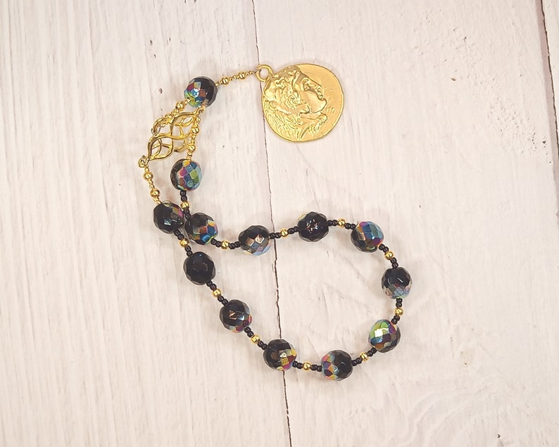 Pode incluir: Um bracelete de contas pretas com um fecho dourado que apresenta o perfil de uma pessoa. As contas s&atilde;o facetadas e t&ecirc;m um brilho iridescente arco-&iacute;ris.