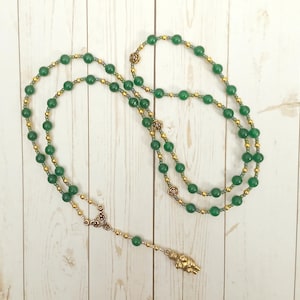 Peut inclure: Un collier de perles vertes et dorées avec un petit pendentif en forme de figurine dorée. Le collier est composé de deux rangs de perles, l'un avec des perles vertes et l'autre avec des perles dorées.