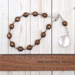 May include: A brown beaded necklace with a silver charm. The necklace is made of faceted beads and has a silver clasp. The charm is a round disc with a smooth surface.