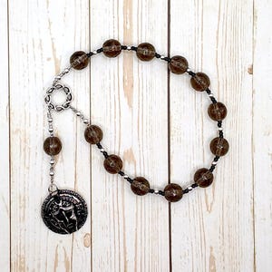 Könnte beinhalten: Ein Rosenkranz mit großen, durchscheinenden braunen Perlen und kleineren schwarzen und weißen Perlen. Der Rosenkranz hat ein silberfarbenes Mittelstück und einen Anhänger mit geprägtem Design. Die Perlen sind auf einer schwarzen Schnur aufgefädelt.
