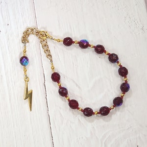 May include: A gold-toned bracelet with faceted, deep red beads and smaller gold beads. A gold lightning bolt charm and a shimmering, iridescent bead dangle from the chain. The bracelet is displayed on a white wooden surface.