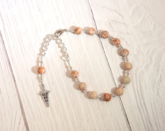 May include: A bracelet made with light brown and white marbled beads and a silver chain with a medical symbol charm.