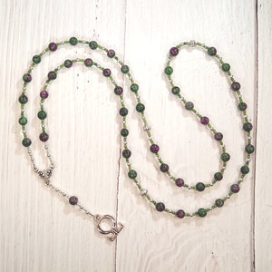 May include: A green and pink gemstone beaded necklace with a silver charm. The necklace is made with small round beads and has a silver clasp.